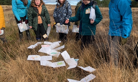 A group of people taking part in an art workshop on moorland - there are sketches of the landscape and botany on the grass in front of them