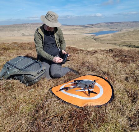 A 4 rotor UAV sits on an orange disk on open moorland; a man uses a control to prepare it for take off