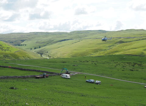 A helicopter with a long cable trailing below it, coming into a high valley to pick up more cargo